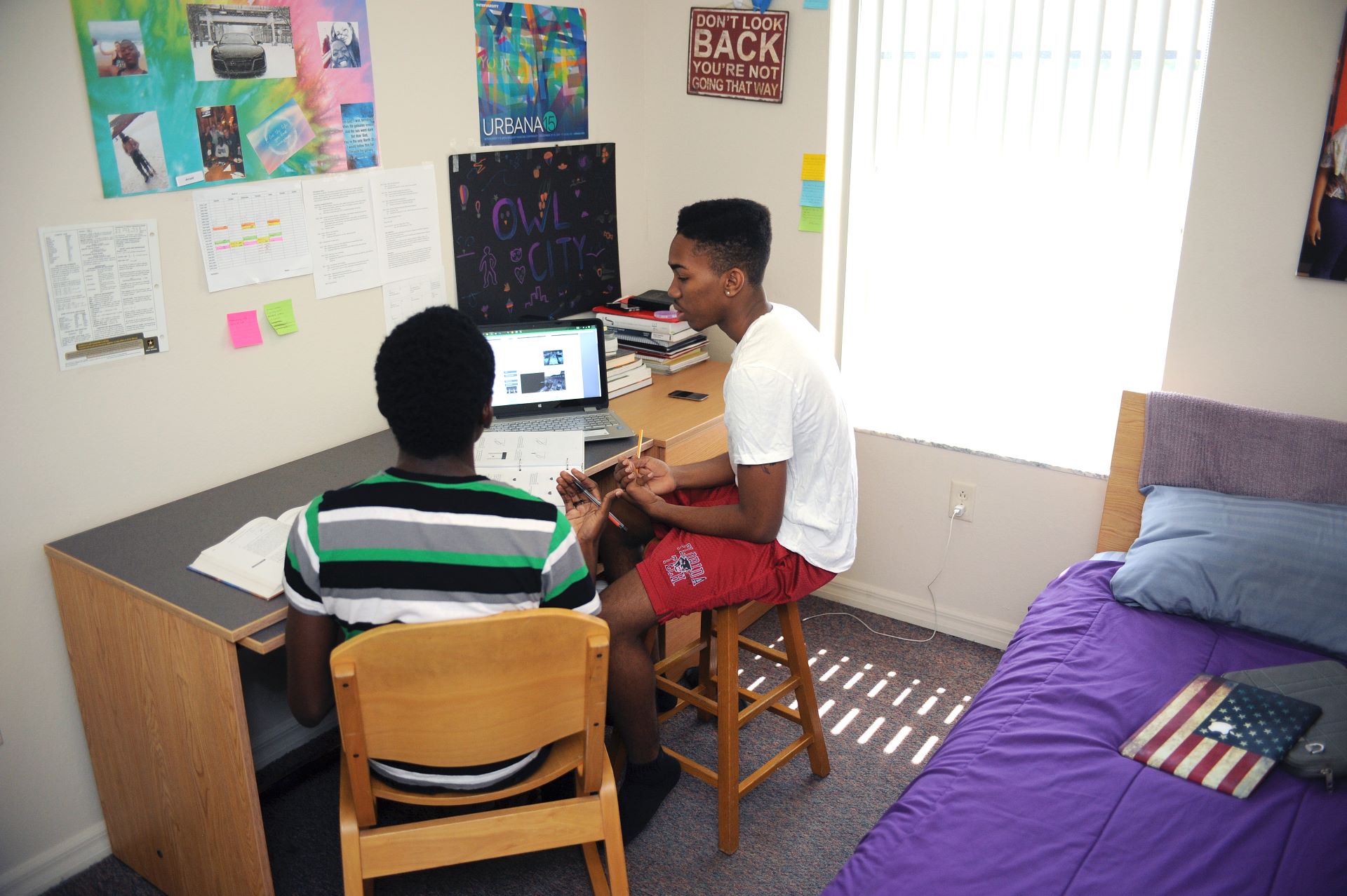 Two students sitting at a desk in a dorm room, one with a laptop open, discussing and working on a project